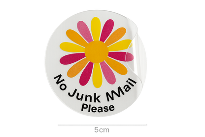 a photograph of a clear round sticker that is 5cm across. The sticker is half peeled off it's white paper backing.  The sticker has a bold daisy flower printed on it in multiple colours, with the text ''No Junk Mail Please''. The sticker is clear.