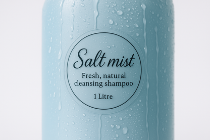 a photograph of a clear round sticker that is 5cm across. The sticker is stuck onto a pale blue shampoo bottle with water dripping down the side of the shampoo bottle. The sticker has a pretty logo printed on it, with the text ''Salt mist Fresh, natural cleansing shampoo, 1Litre''